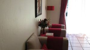 En stue med en beige sofa, et rødt pled, en vase med blomster og et billede på væggen.