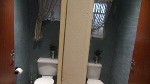 A bathroom with two toilets, a mirror, and a window with curtains. - Cozy Triple Room With King Sized bed and Single Bed, Near Bloemfontein (Bloemfontein)