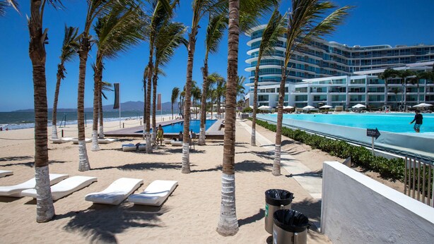 A beachside resort with palm trees, lounge chairs, a swimming pool, and a hotel building in the background. - Studio Ocean View in the Best Location of Vallarta (Nuevo Nayarit)