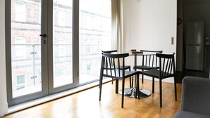 Un apartamento moderno con comedor que incluye una mesa redonda y cuatro sillas, ventanas amplias con vista a un edificio de ladrillo y una cocina con refrigerador.