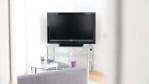 Ein modernes Wohnzimmer mit einem Flachbildfernseher auf einem Glastisch, einem runden Glastisch mit einigen Gegenständen und einem grauen Sofa.
