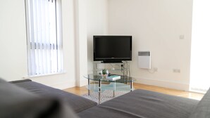 Ein modernes Wohnzimmer mit Flachbildfernseher auf einem Glastisch, einer Couch und einem Fenster mit Jalousien.