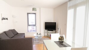 Ein modernes Wohnzimmer mit grauem Sofa, einem Glastisch, einem Flachbildfernseher und einem Esstisch mit Stühlen.