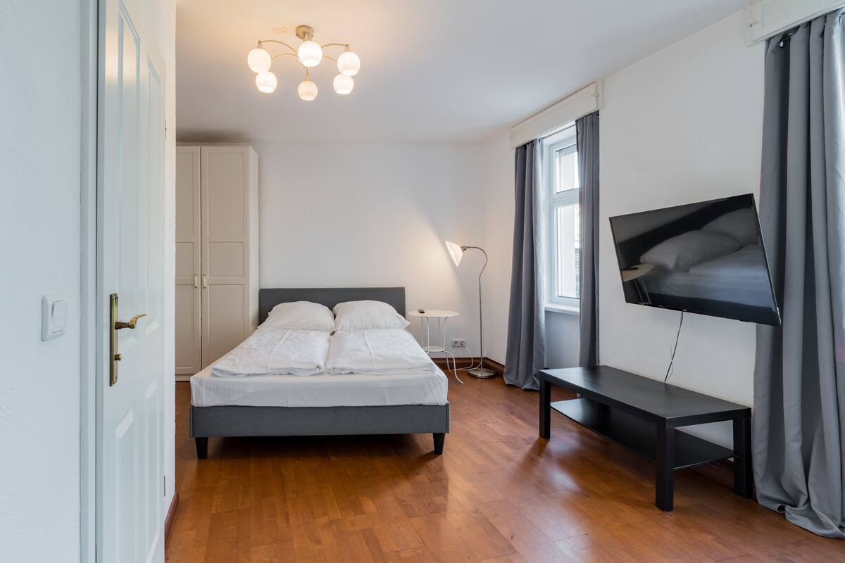 A bedroom with a bed, a TV mounted on the wall, a bedside table, and a window with curtains.