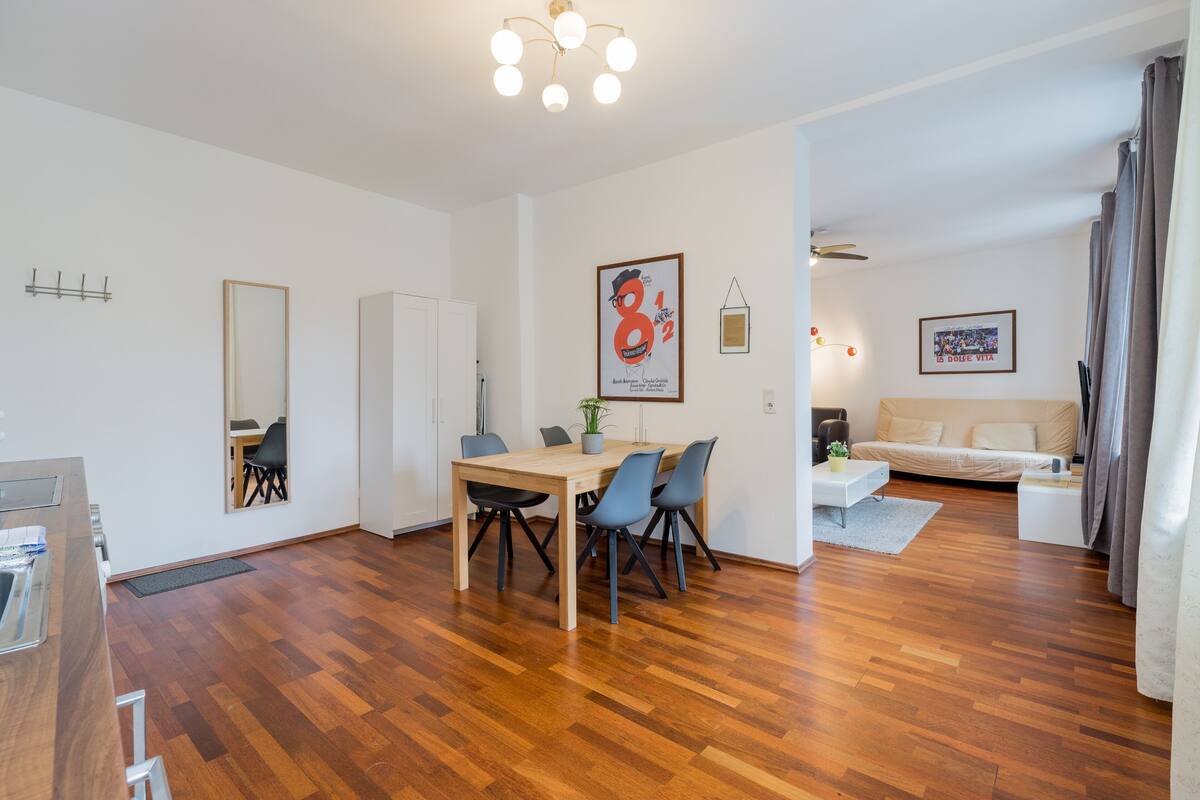 A modern living room with a wooden floor, a dining area with a table and chairs, a sofa, and a TV.