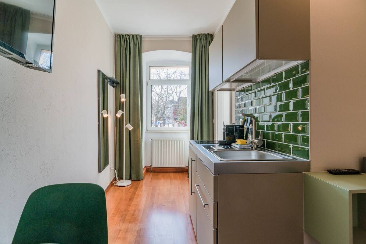 A compact kitchen with a window, green curtains, and a green chair.