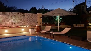 Une piscine entourée de chaises longues et agrémentée d’un parasol, le tout délimité par une clôture blanche illuminée.