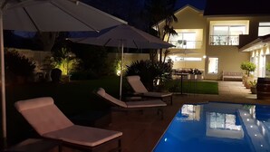 Ein Poolbereich mit Liegestühlen und Sonnenschirmen, ein Essbereich mit Tischen und Stühlen sowie ein zweistöckiges Gebäude mit beleuchteten Fenstern.