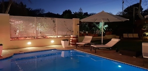Une piscine entourée de chaises longues et agrémentée d’un parasol, le tout délimité par une clôture blanche illuminée.