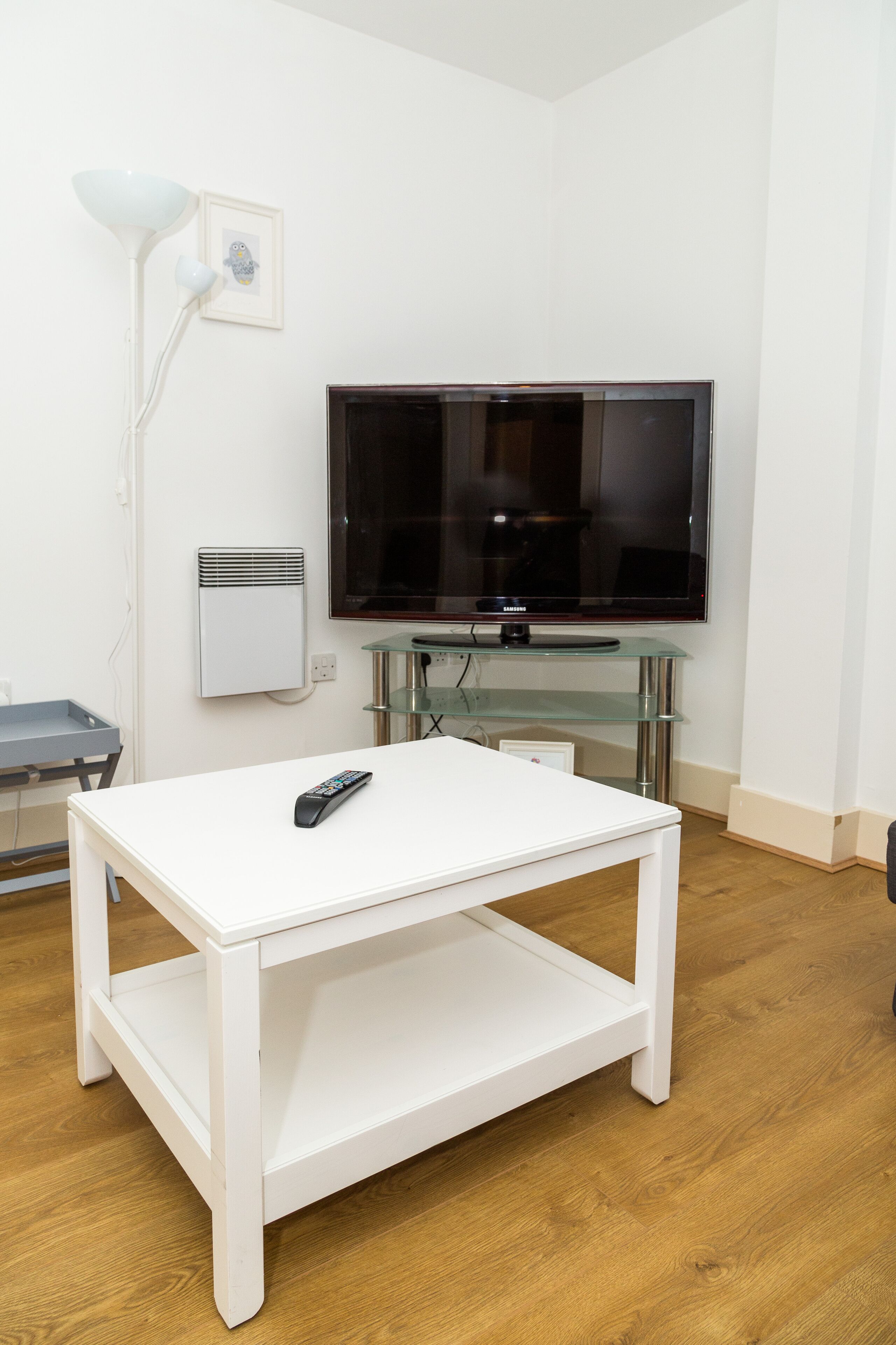A white table with a remote control, a flat-screen TV on a glass stand, a white wall, and a wooden floor.