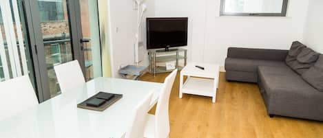 A modern living room with a glass dining table, white chairs, a grey sofa, a flat-screen TV, and a small white side table.