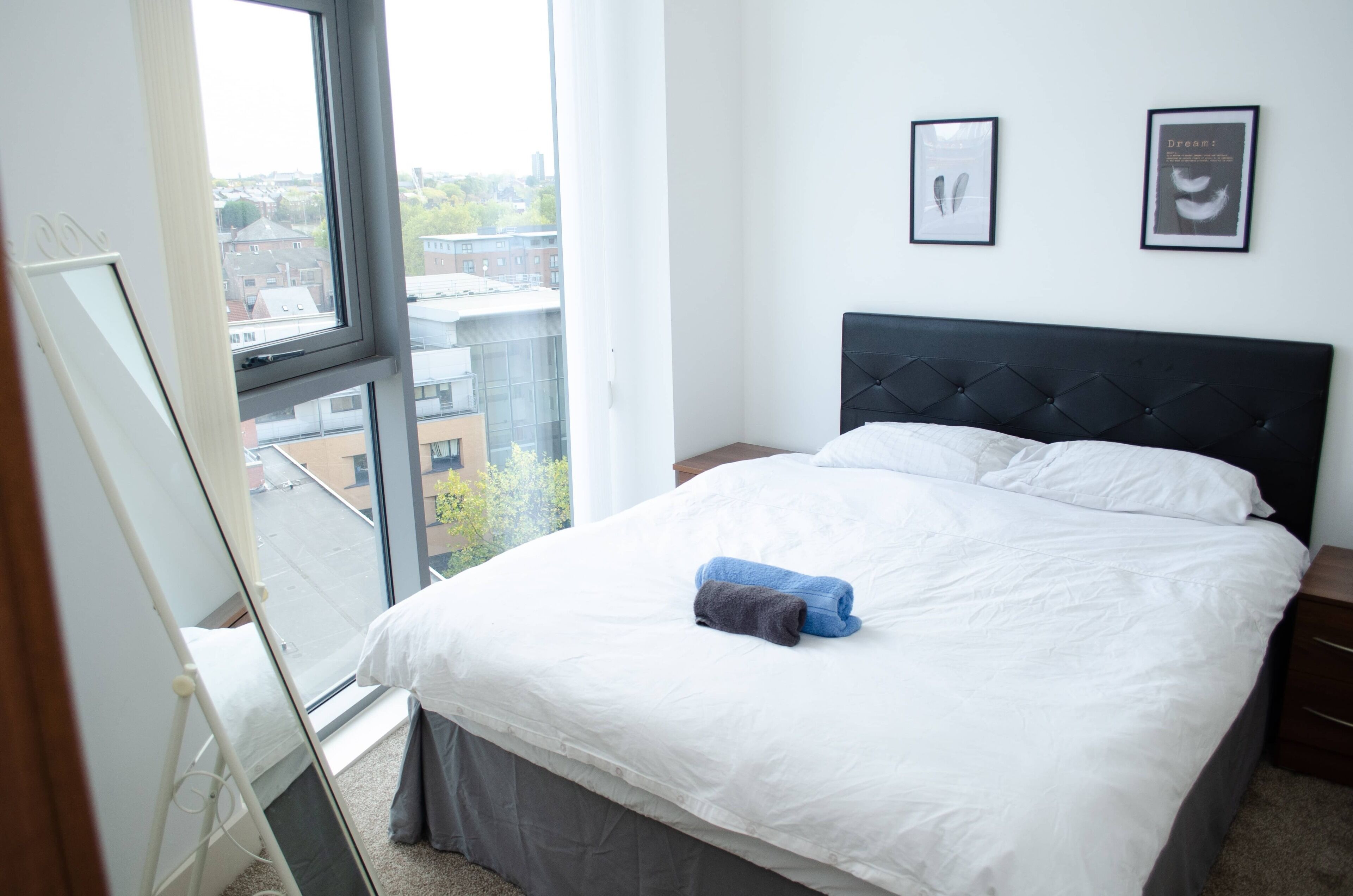 Habitación de hotel con un ventanal grande, una cama con ropa blanca, un cabecero oscuro y dos cuadros en la pared.