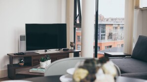 A modern living room with a flat-screen TV, a small table with a bowl of fruit, a sofa, and a view of buildings through large windows. - Kent Street - Apt 16 (Liverpool)