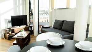 A modern living room with a sofa, a TV, a dining table with white bowls, and a view of buildings through large windows. - Kent Street - Apt 16 (Liverpool)