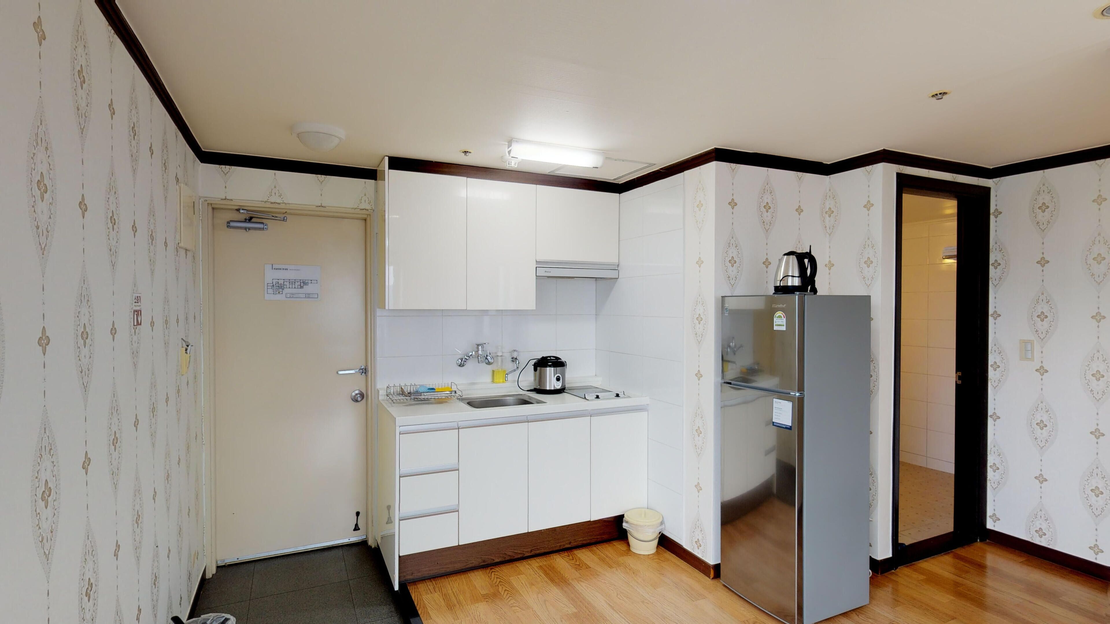 A compact kitchen with white cabinets, a stainless steel refrigerator, a kettle, and a sink.