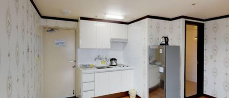 A compact kitchen with white cabinets, a stainless steel refrigerator, a kettle, and a sink.