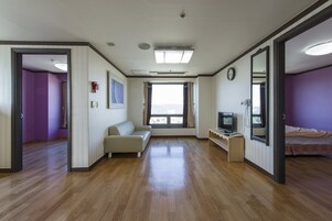 Una habitación con piso de madera, un sofá, un televisor y una cama. Hay ventanas y una puerta.