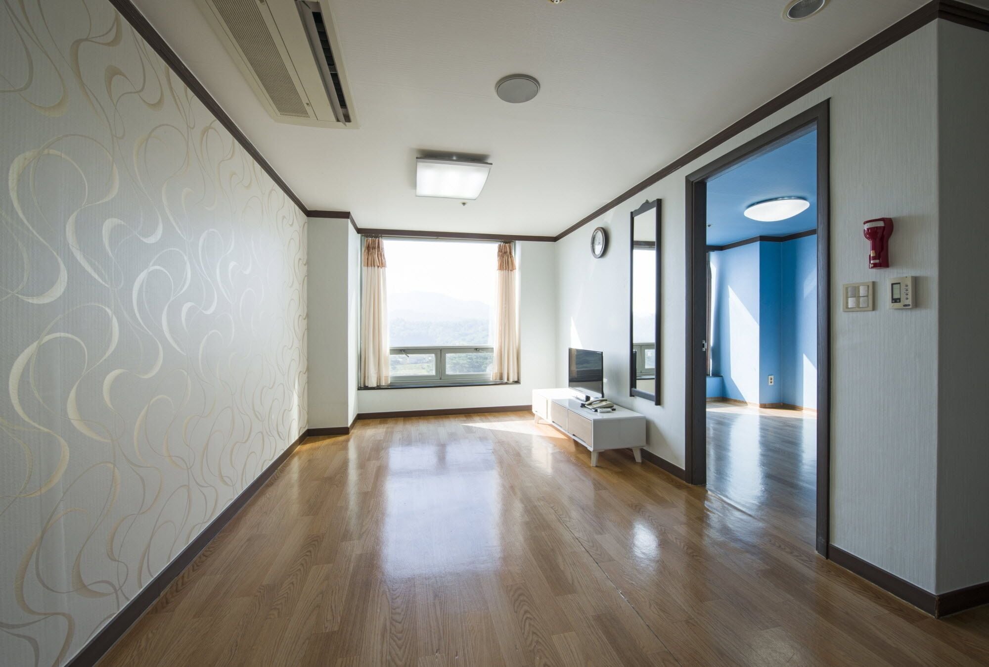 A hotel corridor with wooden flooring, a white wall with a wavy pattern, a window with curtains, a white console table with a television, and a blue wall with a door.