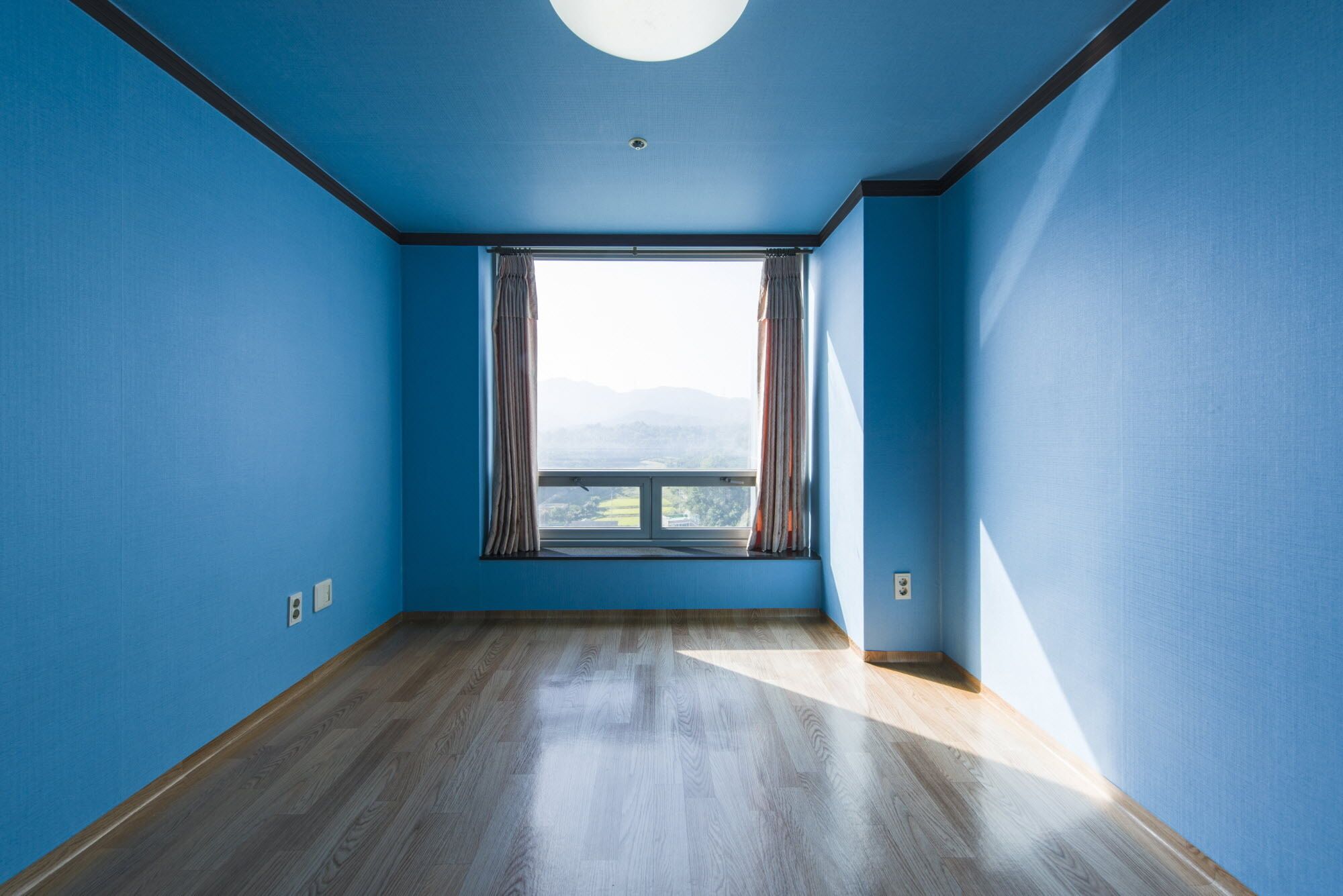 A room with blue walls, a wooden floor, and a large window with curtains.