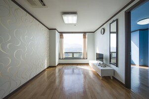 Una habitación de hotel con piso de madera, un ventanal con cortinas, una mesita de noche, un televisor y un reloj en la pared.