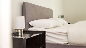 Cama bem arrumada com roupa de cama branca e cabeceira cinzenta, mesa de cabeceira com abajur e uma pequena cômoda.