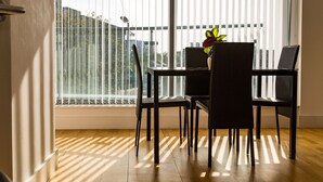 Uma área de refeições com mesa e cadeiras, um vaso de planta e vista através de grandes janelas.
