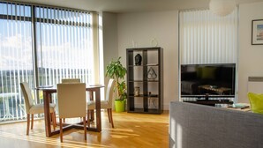 A modern living room with a dining area, a large window with blinds, a flat-screen TV, and a bookshelf. - Kent Street - Apt 8 (Liverpool)