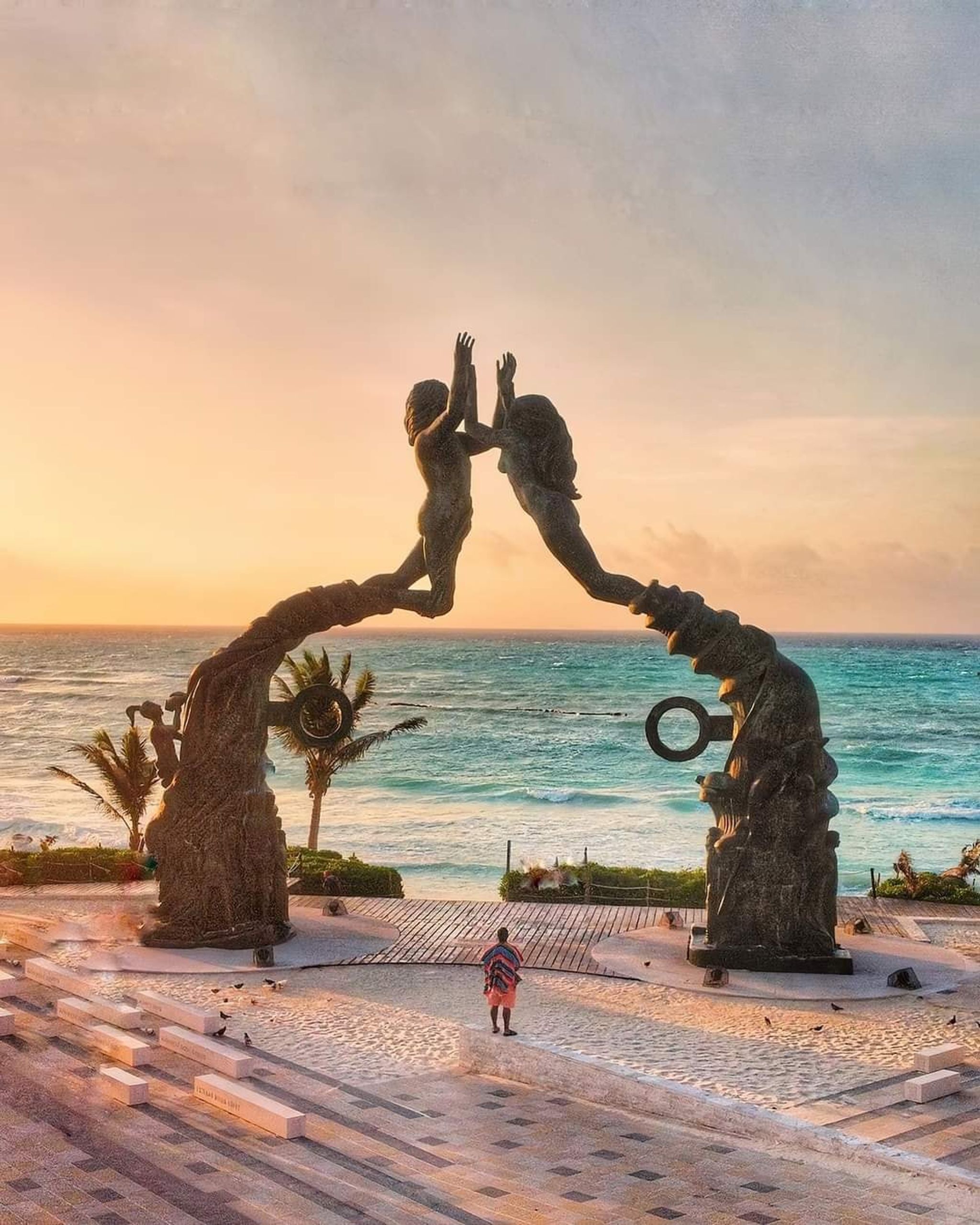 Escultura de dos figuras extendiéndose la una hacia la otra, con el atardecer en una playa de fondo.