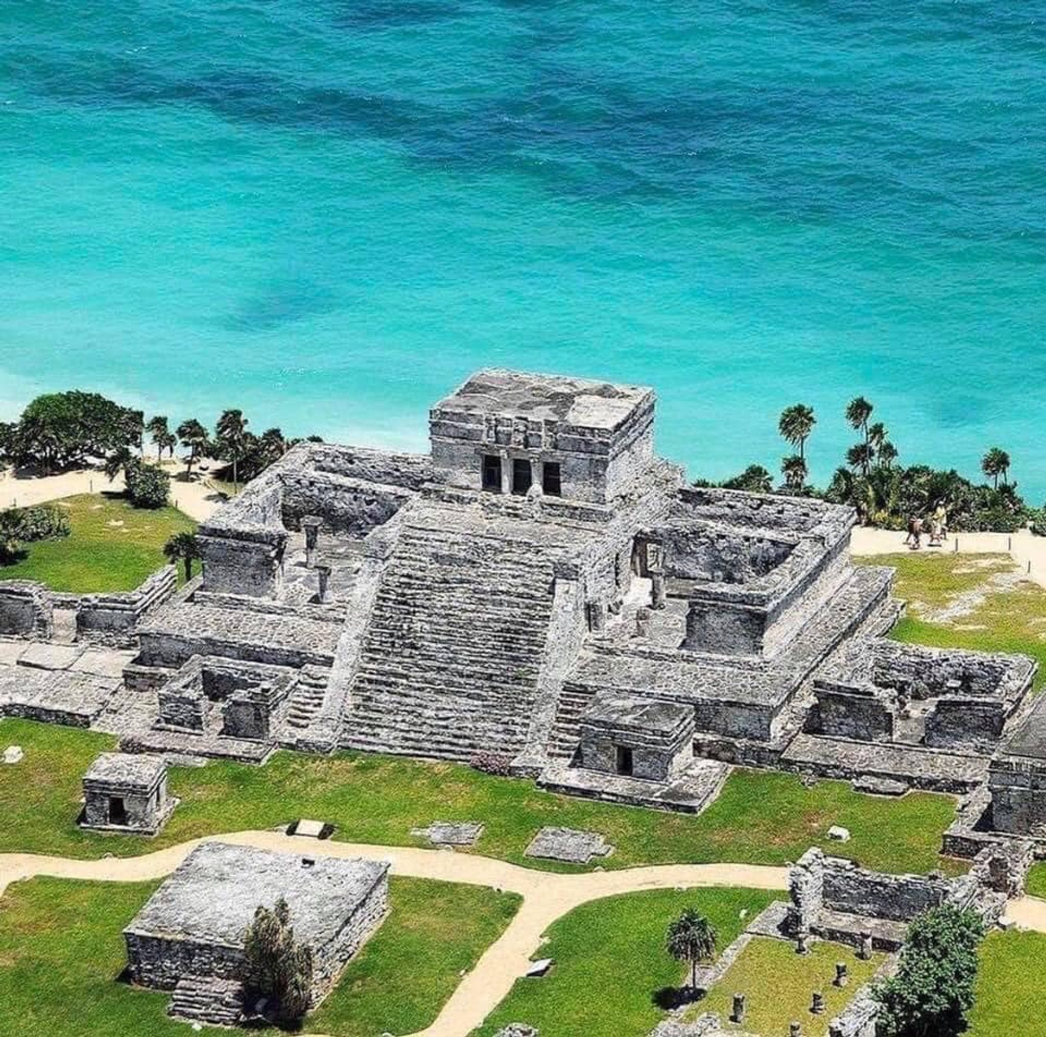 An aerial view of a large ancient Mayan pyramid complex with multiple structures, surrounded by lush greenery and the ocean.