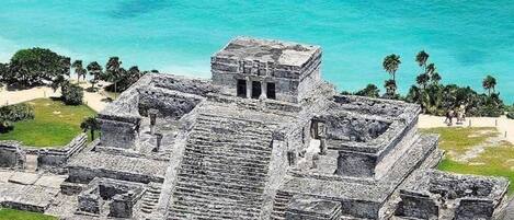 An aerial view of a large ancient Mayan pyramid complex with multiple structures, surrounded by lush greenery and the ocean.