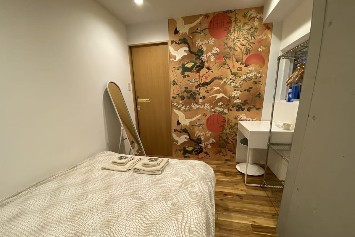 Un dormitorio con cama, una puerta de madera, un muro con arte tradicional, una mesita de noche blanca y un baño pequeño con espejo y lavamanos.