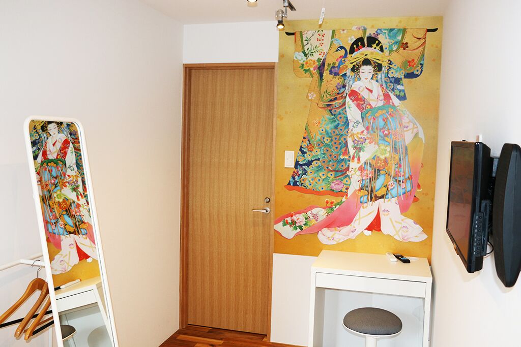 A room with a traditional Japanese painting on the wall, a wooden door, a white desk with a chair, and a television mounted on the wall.