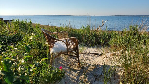 En flätstol med en vit kudde på en sandstrand, med utsikt över ett lugnt vattenområde.