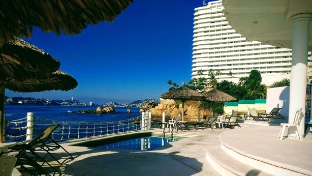 Ein Poolbereich mit Liegestühlen, einem kleinen Schwimmbecken und Blick auf den Ozean sowie ein Hotel im Hintergrund.