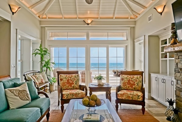 A spacious living room with a view of the ocean, a dining area with a table and chairs, and a kitchen area with cabinets and a stone fireplace.