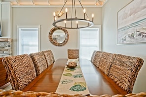 A dining room with a long wooden table, woven upholstered chairs, a central mirror, and a chandelier.