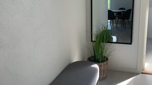 A corner of a room with a grey upholstered bench, a large mirror reflecting a dining area, and a potted plant on the floor. - Just Sleep (Brande)