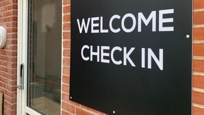 A black sign with white text that reads "WELCOME CHECK IN" mounted on a brick wall next to a door. - Just Sleep (Brande)