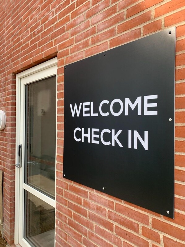 A black sign with white text that reads "WELCOME CHECK IN" mounted on a brick wall next to a door.