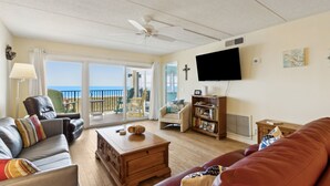 A living room with a sofa, armchair, coffee table, and a TV. There is a bookshelf and a window with a view of the ocean. - Oceanfront Beach Condo with Private Balcony, Short Walk to Pool and Beach by RedAwning (Fernandina Beach)