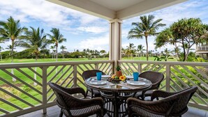 A covered outdoor dining area with wicker chairs and a round table, overlooking a well-maintained lawn and palm trees. - Fairway S Waikoloa J21 2 Bedroom Villa by RedAwning (Waikoloa)