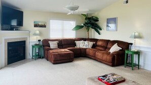 A living room with a sectional sofa, a fireplace, a ceiling fan, and a large plant. - Salt, Sand And Sudds 5 Bedroom Home by RedAwning (Supply)