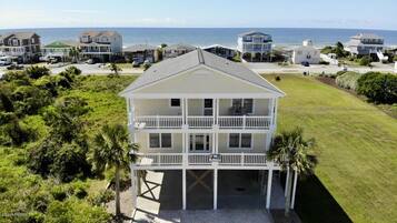 A two-story beachfront house with a balcony, surrounded by lush greenery and a view of the ocean.