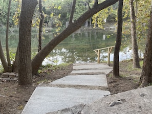 Un sendero de concreto que lleva a un banco de madera junto a un lago tranquilo, rodeado de árboles con hojas otoñales.