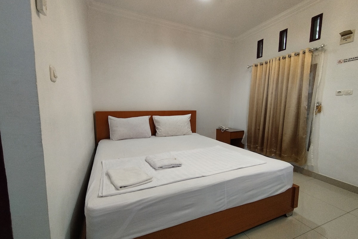 A neatly made bed with white linens and a folded towel, a wooden headboard, a nightstand with a lamp, and a window with curtains.