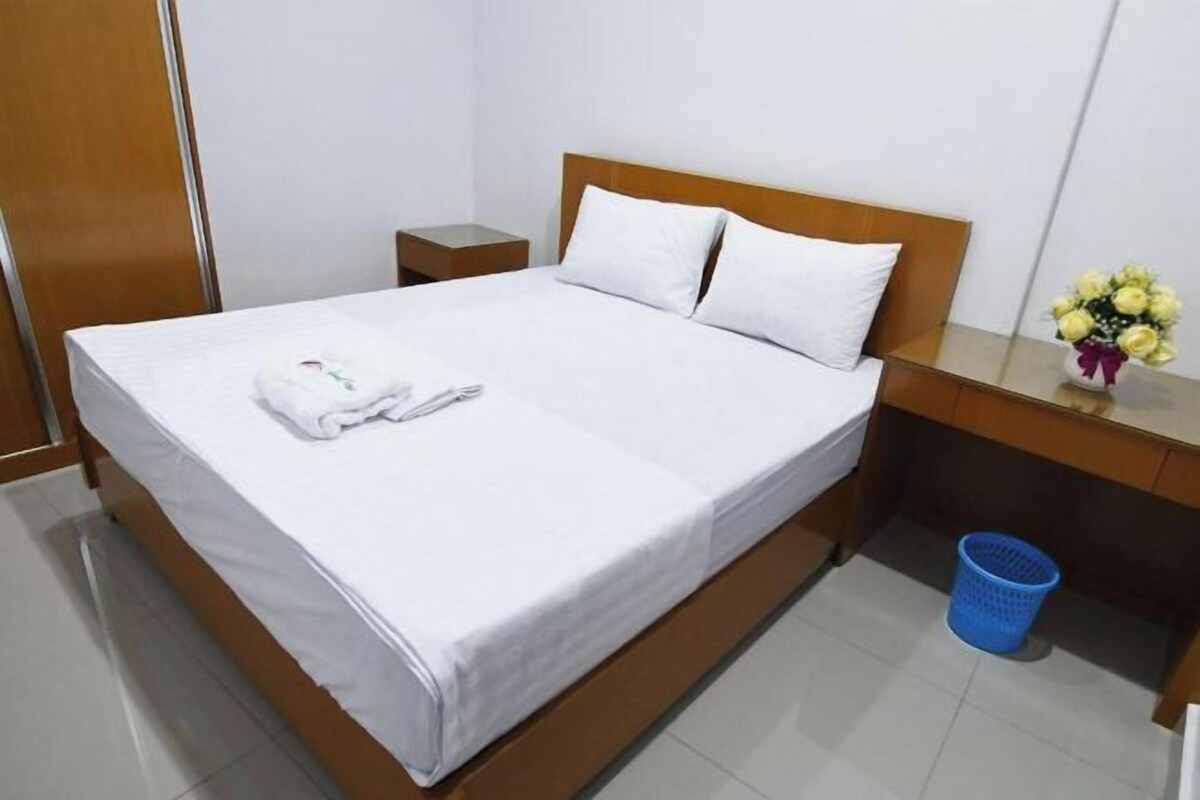 A neatly made bed with white linens, a wooden headboard, a small bedside table with a vase of yellow flowers, and a blue laundry basket.