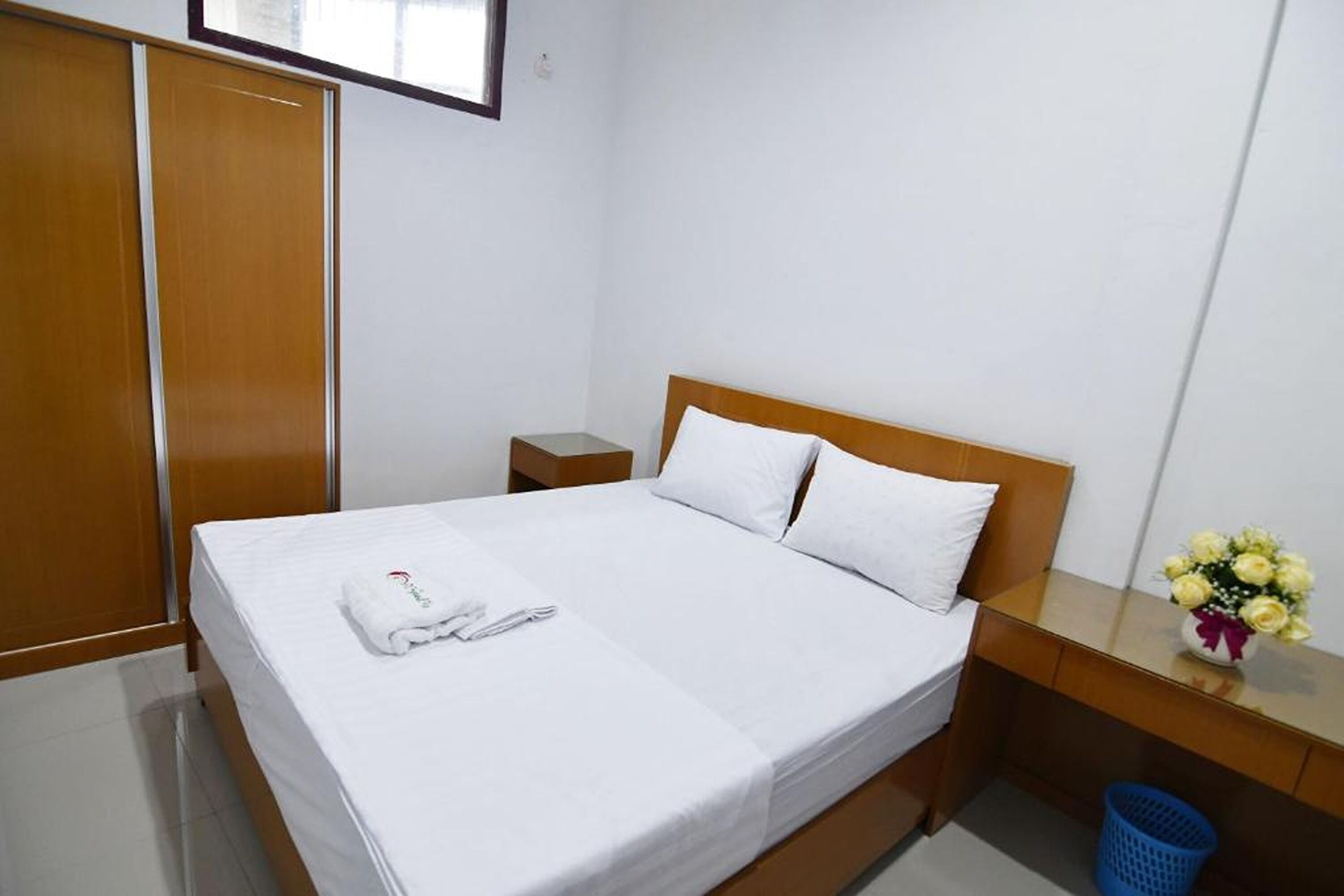 A hotel room with a bed, a nightstand, a wooden wardrobe, and a desk with a vase of flowers.