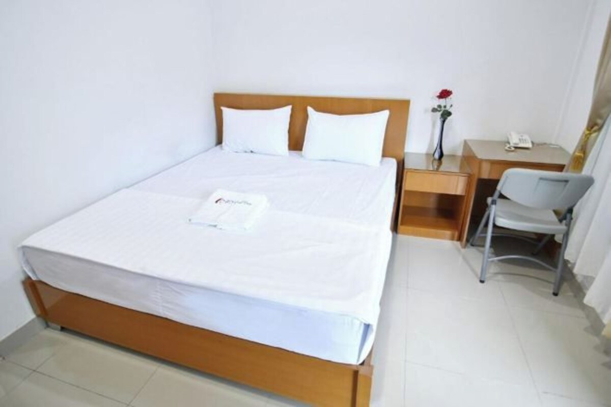A neatly made bed with white bedding and pillows, a wooden headboard, a small bedside table with a phone, and a chair.