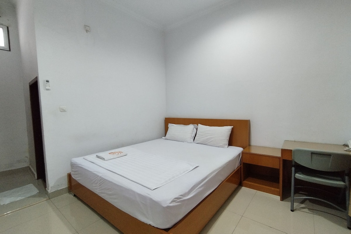 A small, clean room with a single bed, a wooden nightstand, a desk, and a chair.