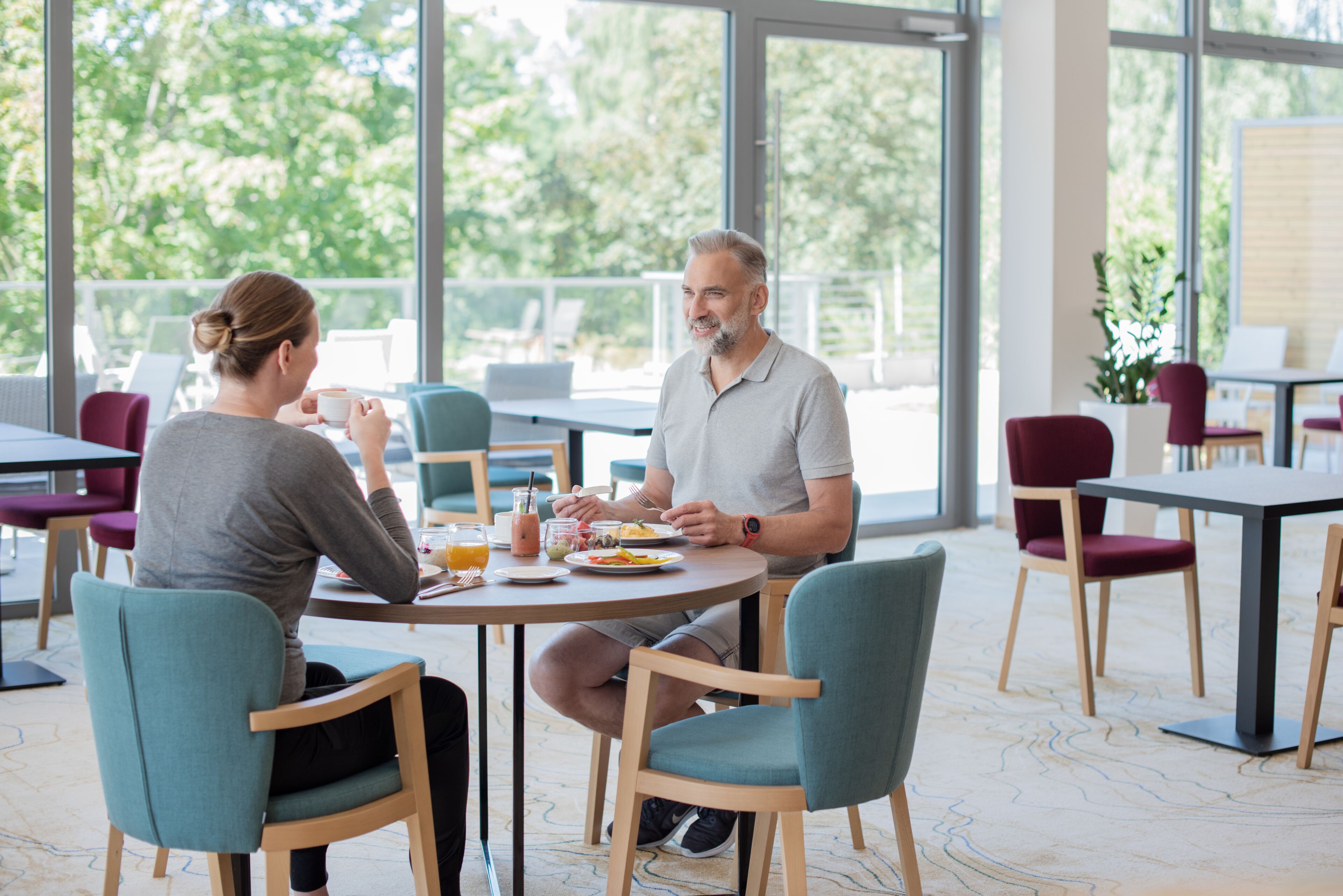 A dining area with round tables and chairs, large windows with a view of greenery, and a casual dining atmosphere.